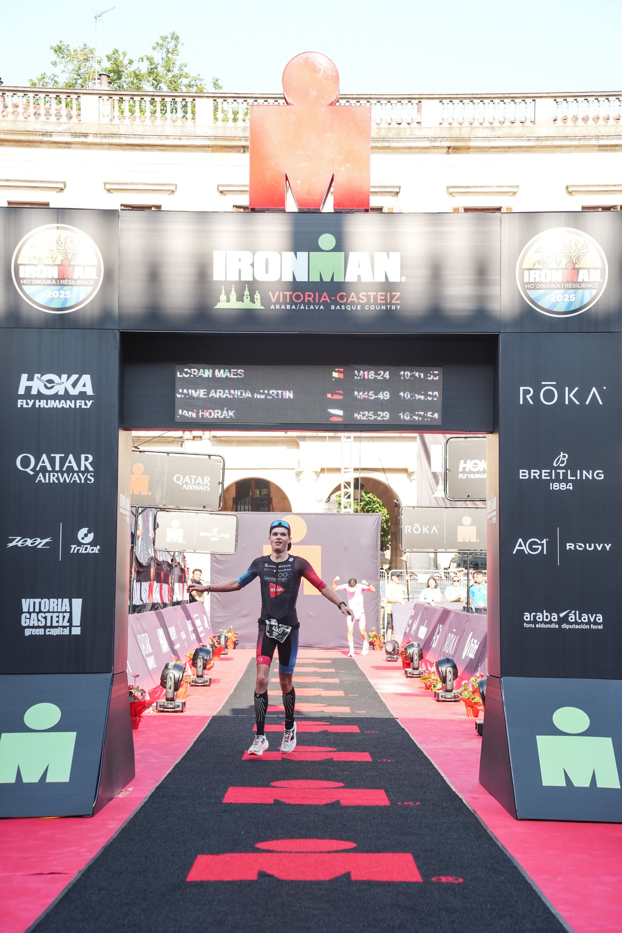 Loran Maes onder de finishboog van IRONMAN Vitoria-Gasteiz, zijn naam bovenaan het scorebord